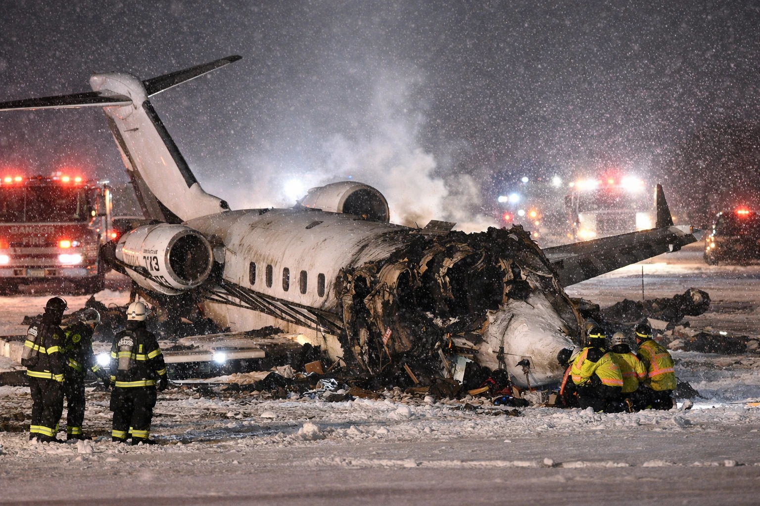 Private jet crash scene at Bangor International Airport in Maine during winter snowstorm, with emergency responders working on the wreckage.